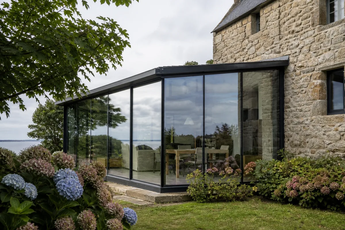 Structure vitrée adossée à une maison en granit, vue depuis le jardin sous une lumière d&rsquo;après-midi