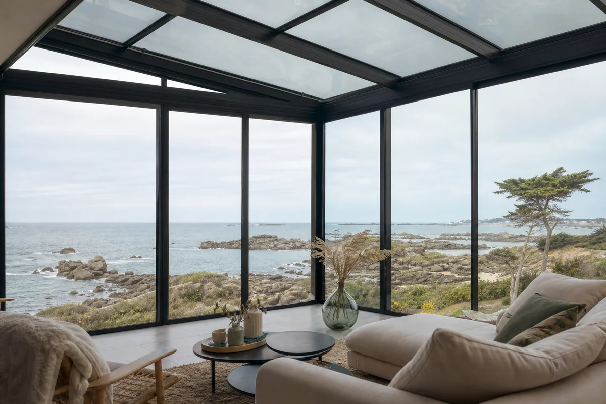 Véranda contemporaine face à la mer sur la côte d’Émeraude, structure aluminium sombre et vue sur les rochers de granit